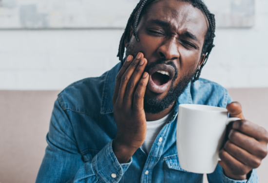 A with a cup of coffee in his left hand while holding the side of his mouth with the right hand, showing signs of discomfort from a toothache.